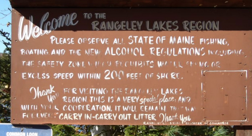 A sign reads, "Welcome to the Rangeley Lakes Region," along with other text, much of which is unreadable due to wear. 